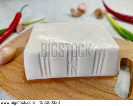 White Cube Of Silken Tofu On The Cutting Board With Chilli Pepper, Onion, And Garlic In The Backgrou