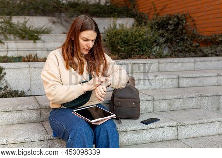 Woman Looking At Clock An Waiting Something. Cheerful Young Femile Sitting On Stairs On City Street,
