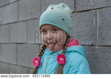 A Portrait Of A Child Against A Gray Stone Wall. A Five-year-old Girl With Her Tongue Out. A Squirmi