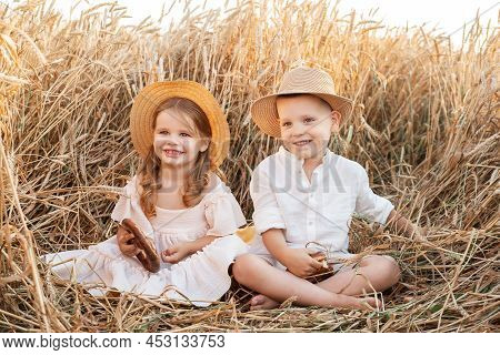 Happy Child Boy And Girl In Straw Hats Sitting In Wheat Field, Eating Bread On Summer. Cottage Core