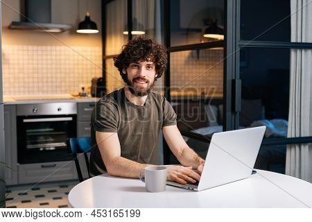 Portrait Of Cheerful Bearded Young Man Using Typing On Laptop Sitting At Table In Kitchen Room With 
