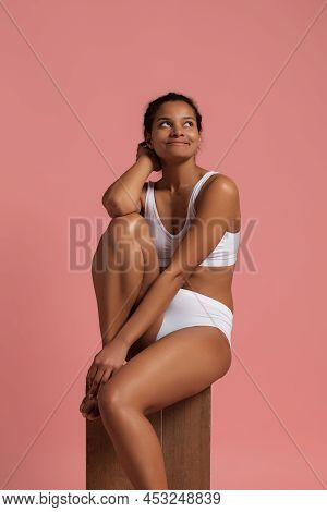 Portrait Of Young Beautiful Slim Woman Sitting, Posing In White Cotton Underwear Isolated Over Pink 
