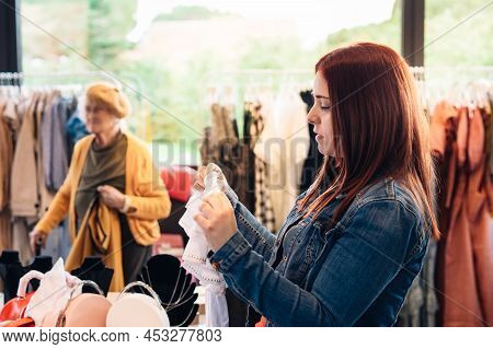 Young Woman Red Hair Looking At Clothes In A Fashion Shop, Deciding What To Buy, Shopping Concept. C