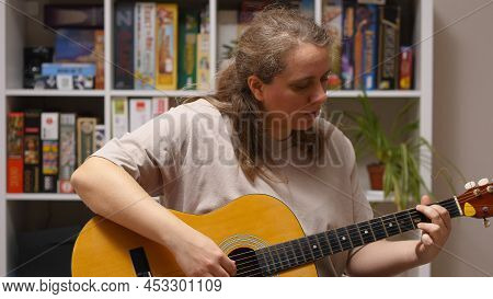 A Girl Plays An Overdub On An Acoustic Guitar Sitting In A Room. Behind Her Is A Rack Of Board Games