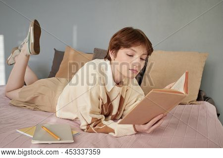 Serious Short-haired Girl In Students Uniform Lying On Bed And Reading Textbook While Prepaing For C