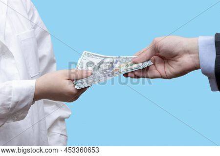 Transfer Of A Bundle Of 100 Dollar Bills To A Female Doctor On A Blue Background. Concept: A Bribe T