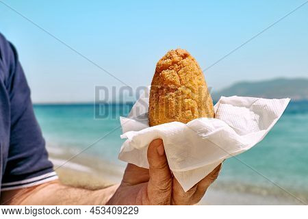 Typical Sicilian Street Food On Seaside Background. Hand Of Man Holding Hot Palatable Arancina (deep