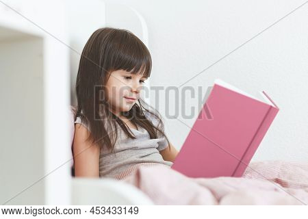 One Todder Girl 5 Years Old Brunette With Long Hair Sits In Bed And Reads A Book.