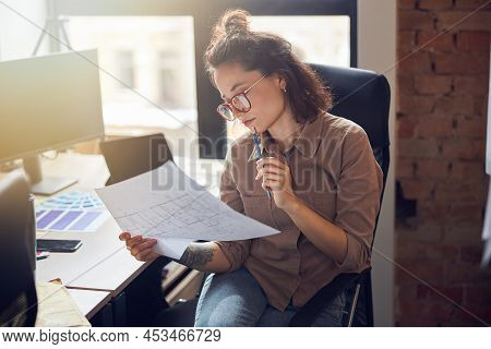 Focused Young Woman Designer Holding And Studying Blueprint Paper While Working On New Interior Desi