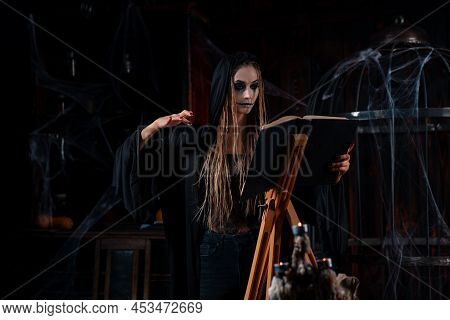 Halloween Concept. Witch Dressed Black Hood Standing Dark Dungeon Room Use Magic Book For Ritual Dev