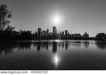 Black And White Photo Of A Lake With Buildings On Background, And The Sun Shining Above Of The City 