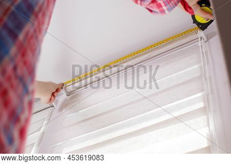 A Handywoman With A Tape Measure Installs White Roller Blinds Indoors. Shutters Day And Night.