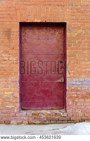 The Old Door Is Inhabited By Tinplate And Painted In Burgundy Color In A Brick Wall.