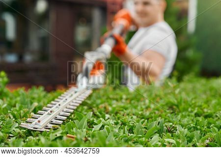 Blur Background Of Male Gardener In Uniform Using Electric Trimming Machine For Pruning Bushes On Ba
