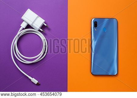 A Modern Blue Phone On An Orange Background And A Charger On A Purple Background.