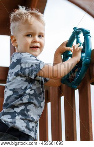 Cute Child Is Driving A Ship Wheel In A Playground. Kid Wearing Camouflage Feels Wild In A Wooden Pl