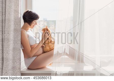 Pregnant Woman In White Underwear With Cute Ginger Cat. Woman In Lingerie Expecting Baby. Risk Of In