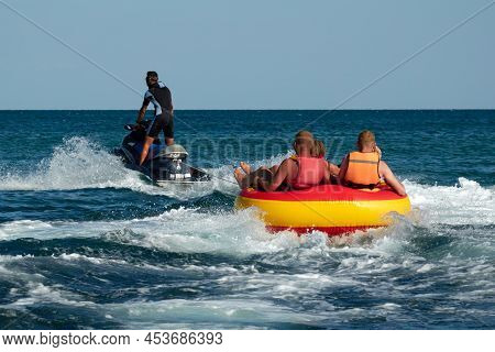 Water Attraction. A Group Of Tourists In Life Jackets Ride On A Water Tubing Towed By A Jet Ski.