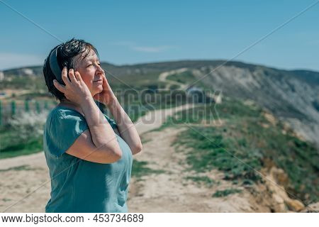Portrait Adult Woman Listens To Music In Headphones In Summer, Spring Against The Backdrop Of Mounta