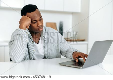 Bored Frustrated African American Guy, Freelancer, Designer, Sitting At A Table At Home, Dressed In 