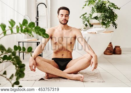 Young Shirtless Man Sitting On Carpet In Lotus Position And Meditating With His Eyes Closed In Bathr