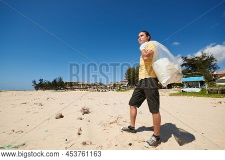 Young Man Standing On Sandy Beach With Bag Of Garbage He Collected