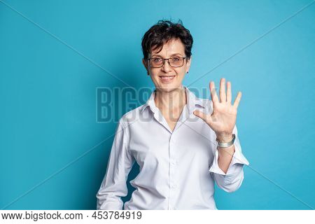 Portrait Of Smiling Middle Adged Woman In Glasses Showing Five Fingers Against Blue Studio Wall Back