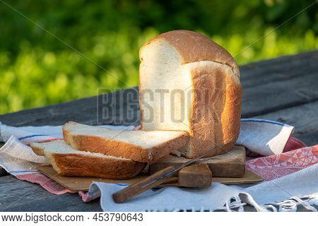 A Freshly Baked Loaf Of White Wheat Bread On A Wooden Table In The Garden. Thick Slices Of Bread Cut