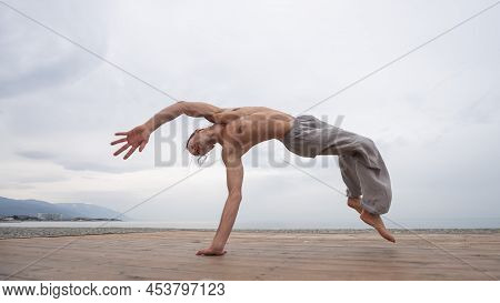 Shirtless Caucasian Man Doing Backflip On Pebble Beach.