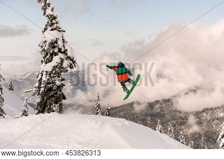 Snowboarder Freerider Jumping From Snow-capped Mountain Slope On Background Of Trees And Cloudy Sky