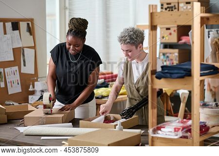 Happy Black Woman Sealing Packed Box With Ordered Goods With Cellotape While Standing Next To Collea