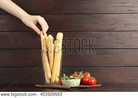 Woman Taking Fresh Delicious Grissino From Glass At Wooden Table, Closeup