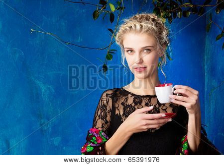 Young beautiful woman with blond hair in braids romantic hairstyle enjoying cup of coffee against blue painted grunge wall with space for text