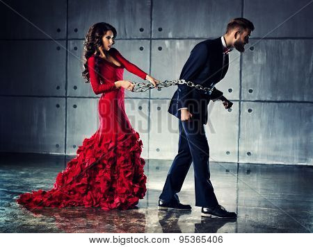 Young woman in red dress holding man on heavy chain. He tries to escape. Elegant evening clothing.