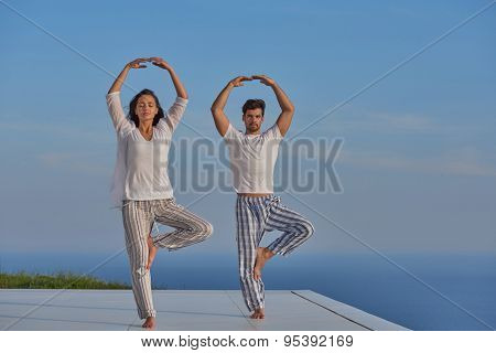 young couple practicing yoga at sunset in modern home terace with ocean and sunset in background
