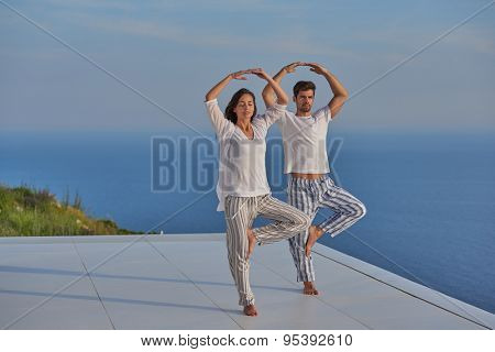 young couple practicing yoga at sunset in modern home terace with ocean and sunset in background