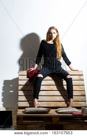 Pretty girl or beautiful woman cute sexy fashion model with long hair in casual clothes sits on backrest of wooden pallet sofa on white wall