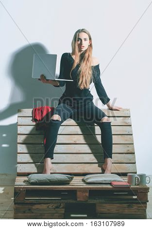 Pretty girl or beautiful woman cute sexy fashion model with long hair in casual clothes with laptop computer sits on backrest of wooden pallet sofa on white wall