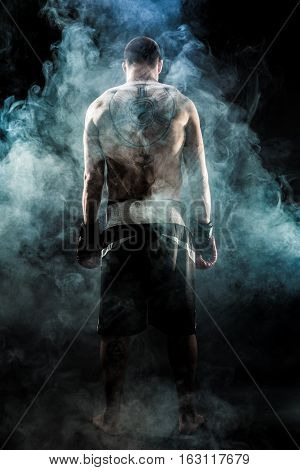 Muscular kickbox or muay thai fighter with tattoo standing back in smoke.