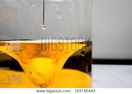 Chemical reaction - A colorless drop of liquid reactant from a dosing needle point is poised to be administered into the vortex of a swirling yellow liquid. Closeup view.