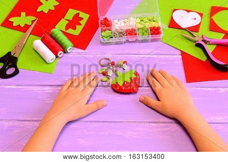 Child made a strawberry keychain of felt and beads. Child shows keychain crafts. Kids hands on a wooden table. Handicraft materials and tools. Kids summer fun crafts