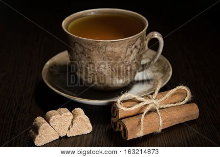 Brown tea cup and saucer, a bundle of cinnamon and sugar on dark wooden background.