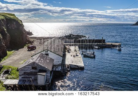 Twillingate, Newfoundland, man works on the engine of one of 4 boats tied up to a wooden, cliff-side dock house for the day, bright sunshine on calm coastal water.