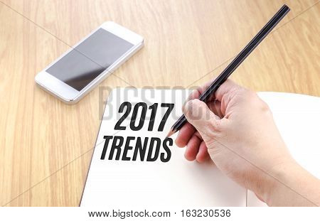 Hand Holding Black Pencil And Writing 2017 Trends On Blank Open Notebook With Smartphone Beside It O
