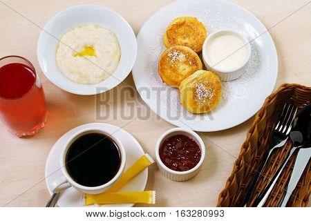 Table set for breakfast with rice porridge, cheesecakes with jam, berry drink, black coffee and cuttlery in wooden container in cafe