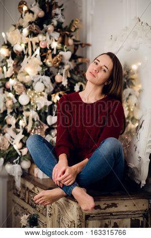 Woman with a Christmas Gift sitting on acncient commode decorated for xmas. Fir tree at background.