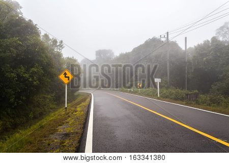 Windy forest road with a curve sign on a foggy day at Doi Inthanon national park Thailand