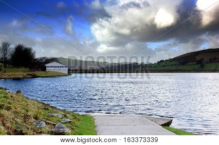 A boat ramp going into the lake with dramatic clouds, also visible a dam and a sail club house