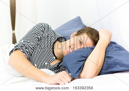 Happy handsome man pretend that he is sleeping in bed alone and smiling for camera. Young man lying on pillow and resting on weekends.
