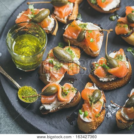 Homemade salmon crostini with cream-cheese, watercress, capers and pesto suace in round black slate stone plate over grey background, selective focus, square crop